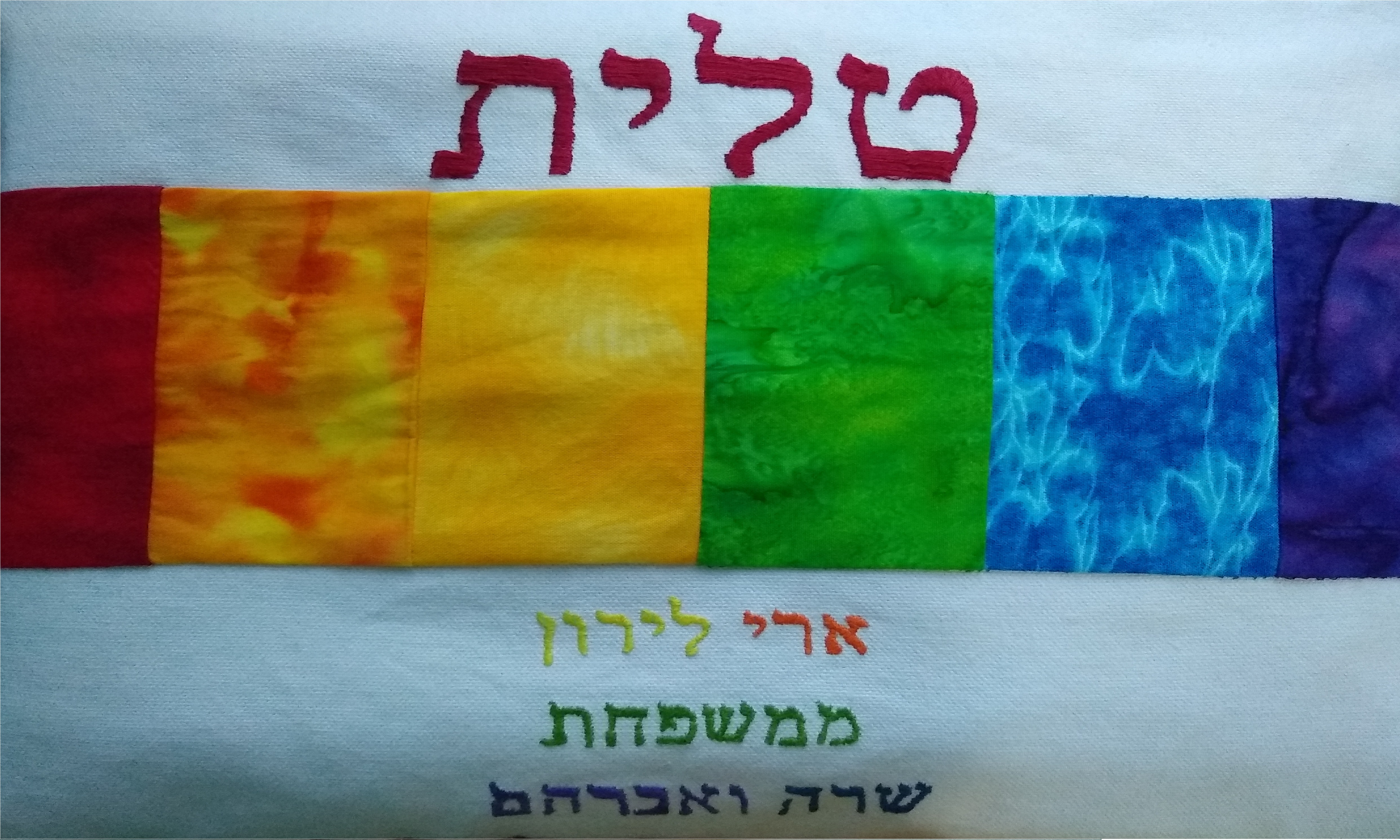 A white tallit bag with rainbow fabric and emboridered hebrew text.