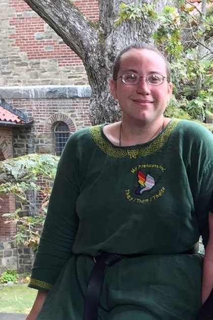 A white person sitting in front of a garden cloister wearing a dark green tunic with gold celtic knotwork embroider at the collar and an embroidered butterfly with the text 'my pronouns are they/them/theirs' worked into the left chest of the garment. Photo.
